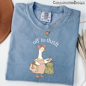 以下が含まれることがあります： 「off to thrift」の文字と、漫画のガチョウのイラストが描かれた、ライトブルーのComfort Colors Tシャツ。ガチョウはリボン、エプロン、ハンドバッグ、ハンガーにかけたドレスを身につけています。金のネックレスがシャツの上にかけられています。