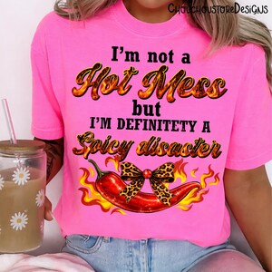 May include: Bright pink t-shirt with the text "I'm not a Hot Mess but I'm definitely a Spicy Disaster." A red chili pepper with a leopard print bow is featured below the text, with flames on either side. The shirt is a casual wear item.