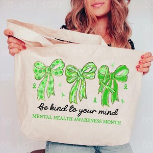 May include: A beige tote bag featuring three green bows with varying patterns and green ribbon accents. The text "Be kind to your mind" and "MENTAL HEALTH AWARENESS MONTH" are printed in black. The bag has a cream-colored strap.