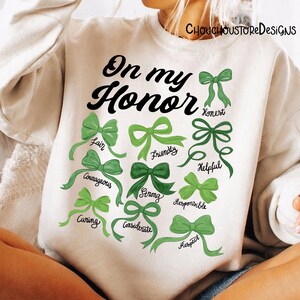May include: White sweatshirt with the text "On my Honor" in black script. Below the text are green bows with words like "Fair", "Courageous", and "Caring". The sweatshirt has the shop name "CHOUCHOUSTORE DESIGNS" printed at the top.