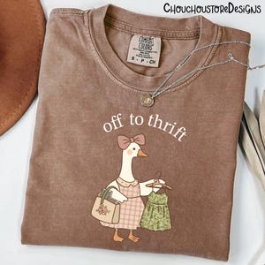 May include: Brown Comfort Colors t-shirt featuring the text "off to thrift" and a cartoon goose with a purse and a dress. The goose wears a pink bow and a plaid dress. A silver necklace is draped across the shirt.
