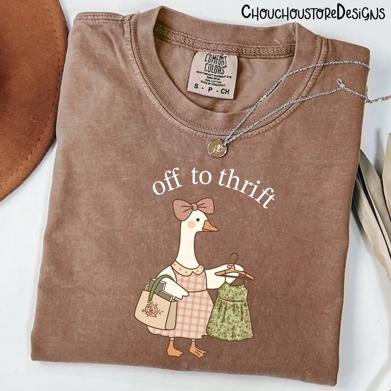 May include: Brown Comfort Colors t-shirt featuring the text "off to thrift" and a cartoon goose with a purse and a dress. The goose wears a pink bow and a plaid dress. A silver necklace is draped across the shirt.