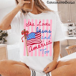 May include: White t-shirt with the text "She Loves Jesus and America Too" in red and blue script. The design includes a cross and an American flag with a pink striped background.