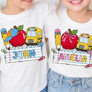 May include: Two white t-shirts featuring a school-themed graphic. The design includes a yellow school bus, a red apple, and a yellow pencil above a piece of lined notebook paper. One shirt displays the name JOHN, and the other displays AMELIA.