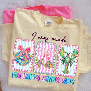 以下が含まれることがあります： 「I was made for happy sunny days」の文字が入った薄い黄色のTシャツ。貝殻、ヒトデと貝殻、ロブスターの3つのフレームグラフィックが特徴です。グラフィックは明るい色です。