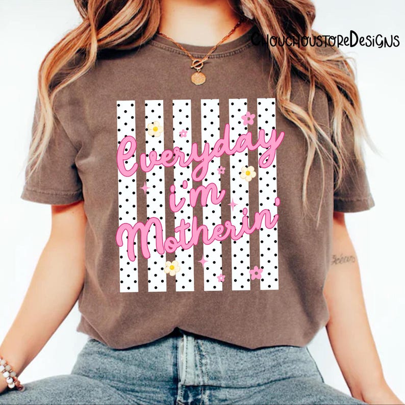 May include: A brown t-shirt featuring the pink text "Everyday I'm Motherin". The design includes white vertical stripes, black polka dots, and small floral details, creating a playful aesthetic.
