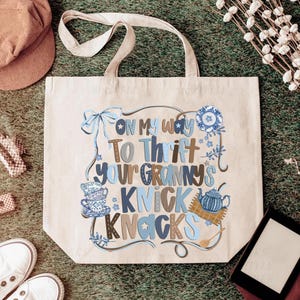 May include: Beige tote bag with the text "On My Way To Thrift Your Granny's Knick Knacks" in blue and brown lettering. The bag features illustrations of teacups, a teapot, and a bow. The bag has a pair of handles.