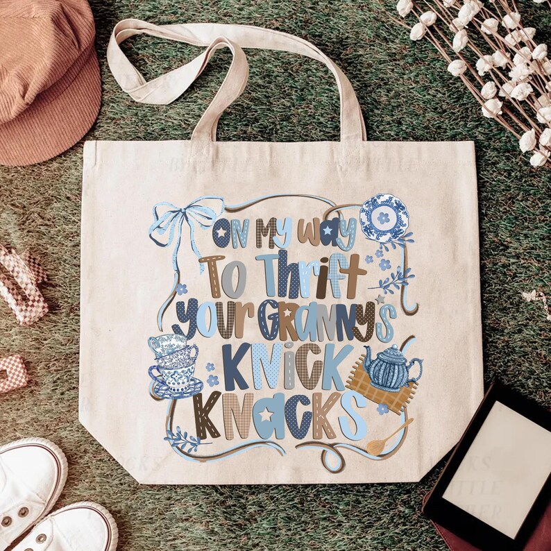 May include: Beige tote bag with the text "On My Way To Thrift Your Granny's Knick Knacks" in blue and brown lettering. The bag features illustrations of teacups, a teapot, and a bow. The bag has a pair of handles.