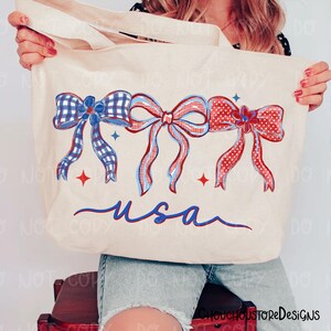 May include: Cream-colored tote bag featuring three decorative bows in red, white, and blue. The bows have plaid and polka dot patterns. The word "usa" is written in blue script below the bows. The bag has cream-colored handles.