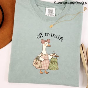 May include: A pale green t-shirt with the words "off to thrift". The shirt features a cartoon goose wearing a bow and apron, holding a purse and a dress. A gold necklace is draped across the shirt.