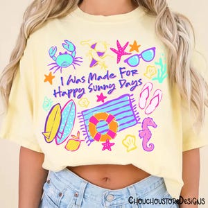 May include: A light yellow t-shirt with a colorful beach-themed graphic. The design includes a crab, sunglasses, starfish, a beach towel, a life preserver, and the text "I Was Made For Happy Sunny Days."