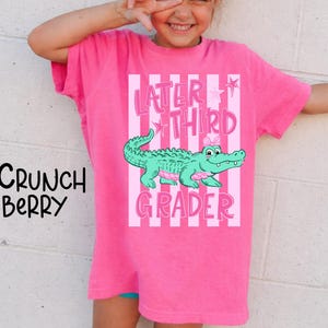 May include: A bright pink t-shirt with the text "Later Third Grader" and a cartoon alligator. The alligator is green with pink accents. The shirt also has vertical pink and white stripes.
