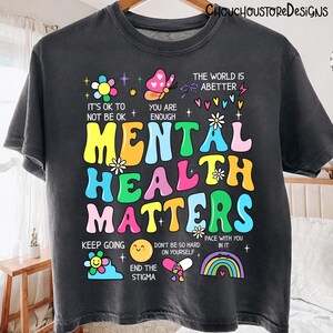May include: Dark gray t-shirt with the words "MENTAL HEALTH MATTERS" in colorful, bubble letters. The shirt includes uplifting phrases like "It's OK to not be OK" and "You are enough," along with various illustrations.