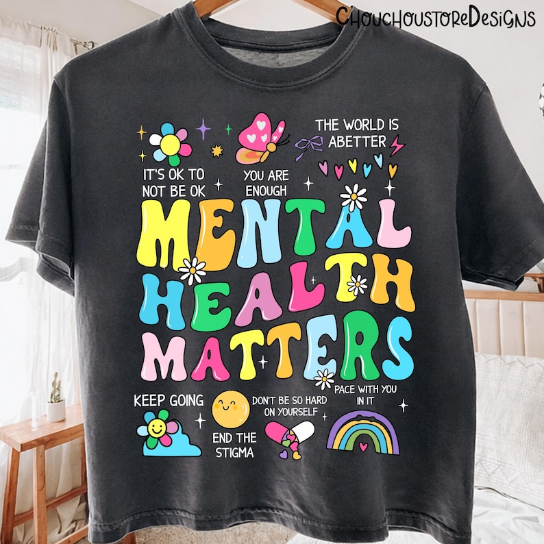 May include: Dark gray t-shirt with the words "MENTAL HEALTH MATTERS" in colorful, bubble letters. The shirt includes uplifting phrases like "It's OK to not be OK" and "You are enough," along with various illustrations.