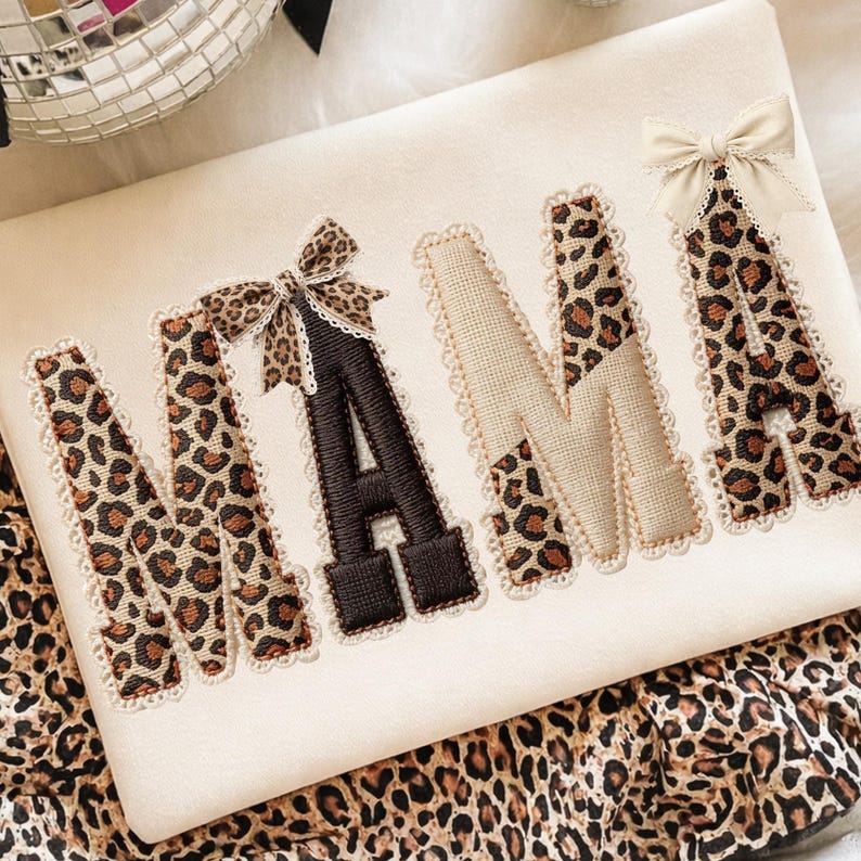 May include: Cream-colored fabric with the word "NANA" embroidered in a mix of leopard print and solid brown. Each letter is outlined with a delicate lace trim and adorned with small bows. The fabric is set against a backdrop of leopard print.