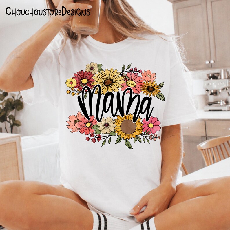May include: White t-shirt featuring the word "Mama" in a black script font, surrounded by a colorful floral arrangement. The flowers include sunflowers, daisies, and other blooms in shades of pink, yellow, and red. The top is a casual style.