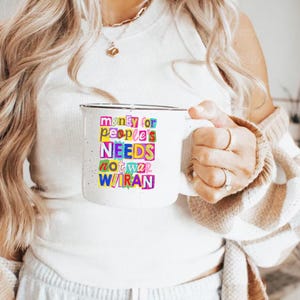 May include: White ceramic mug with a black rim, featuring the text "money for people's NEEDS not war W/IRAN" in colorful, block letters. The mug is held by a person wearing a white top and a beige sweater.