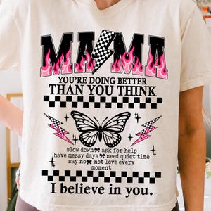 May include: Cream-colored t-shirt with the word "MAMA" in black block letters with pink flame accents. The shirt features a butterfly graphic and the text "YOU'RE DOING BETTER THAN YOU THINK" and "I believe in you."