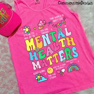May include: Bright pink tank top with the words "MENTAL HEALTH MATTERS" in colorful, bubble letters. The shirt features various illustrations, including flowers, a butterfly, and a rainbow. A pink baseball cap is visible in the upper left corner.