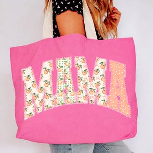 May include: A bright pink tote bag with the word "MAMA" in large, decorative letters. The letters are filled with peach and green gingham patterns. The letter "A" is filled with a peach polka dot pattern. The bag has cream-colored handles.