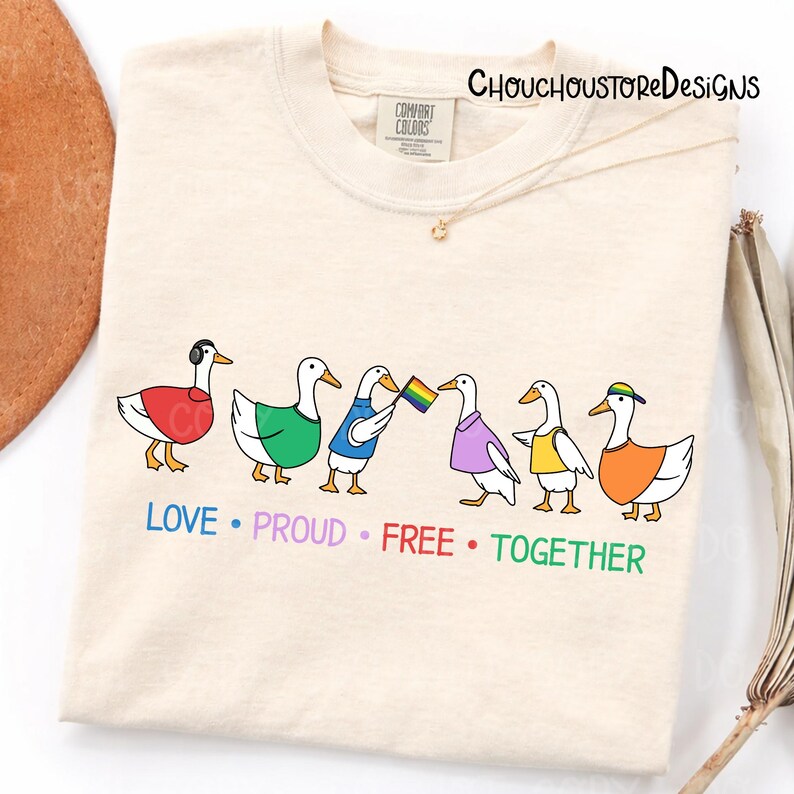 May include: Cream-colored t-shirt featuring a line of cartoon ducks, each wearing different colored outfits and accessories. One duck holds a rainbow flag. The words "LOVE &bull; PROUD &bull; FREE &bull; TOGETHER" are printed below the ducks. The brand name "CHOUCHOUSTOREDESIGNS" is printed at the top.