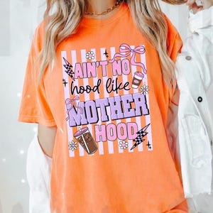 May include: Orange t-shirt with the text "Ain't No Hood Like Motherhood" in a vertical design. The design includes pink bows, flowers, lightning bolts, and coffee cup graphics. The shirt is a short-sleeved, crew neck style.