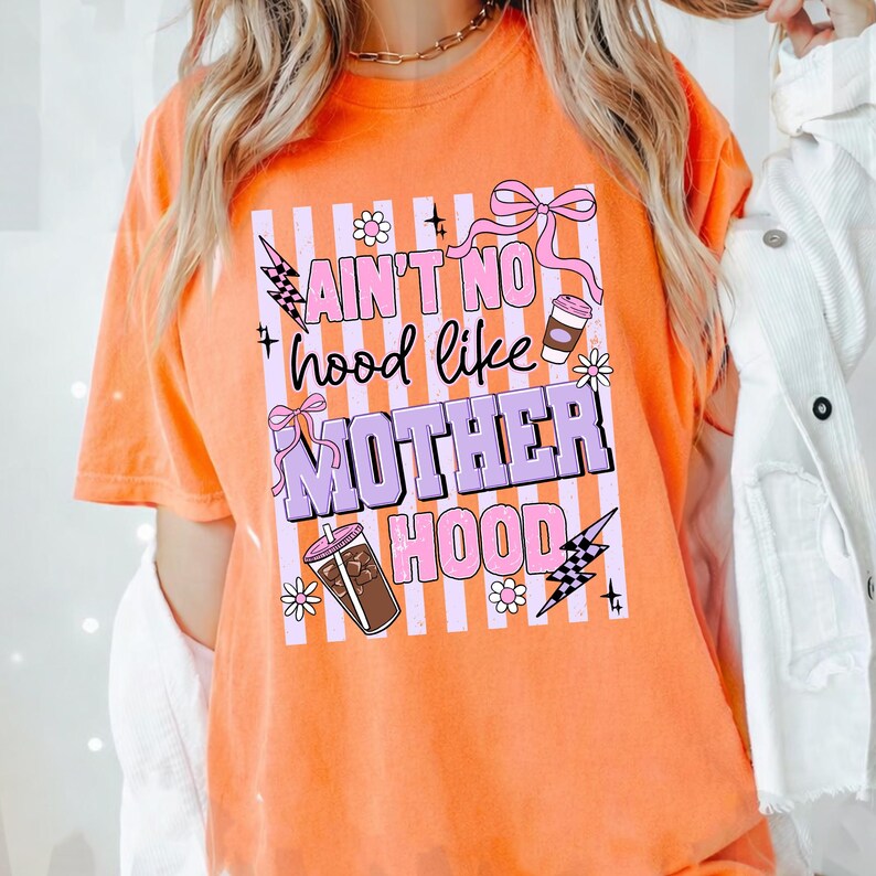 May include: Orange t-shirt with the text "Ain't No Hood Like Motherhood" in a vertical design. The design includes pink bows, flowers, lightning bolts, and coffee cup graphics. The shirt is a short-sleeved, crew neck style.