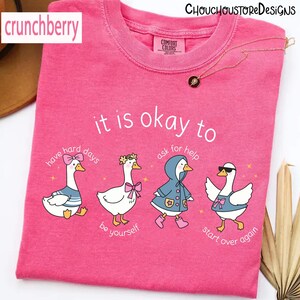 May include: A pink Comfort Colors t-shirt with the text "it is okay to" above four cartoon geese. Each goose is styled differently, with text below each: "have hard days", "be yourself", "ask for help", and "start over again".