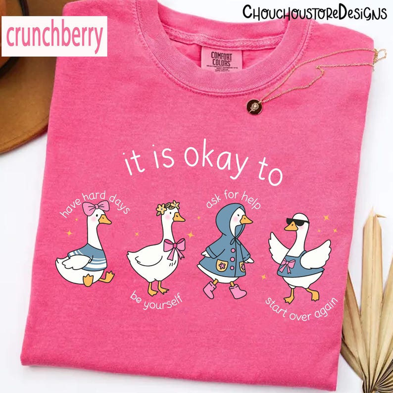 May include: A pink Comfort Colors t-shirt with the text "it is okay to" above four cartoon geese. Each goose is styled differently, with text below each: "have hard days", "be yourself", "ask for help", and "start over again".
