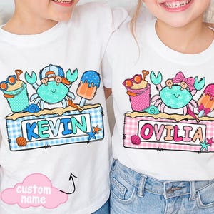 Puede incluir: Camisetas blancas con un gráfico colorido con temática de playa. Una camiseta presenta un cangrejo con un sombrero y el nombre "KEVIN" sobre un fondo a cuadros azul y blanco. La otra camiseta tiene un cangrejo con un lazo y el nombre "OVILIA" sobre un fondo a cuadros rosa y blanco.