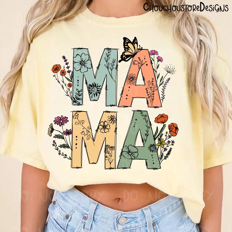 May include: Yellow t-shirt with the word "MAMA" in large, colorful letters. The letters are decorated with floral designs and a butterfly. The shirt is a casual style.