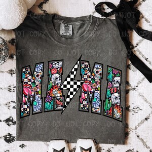 May include: Gray t-shirt with the word "MAMA" in a colorful, graffiti-style design. The letters are filled with skulls, flowers, and other graphics. A black and white checkered lightning bolt is in the center.