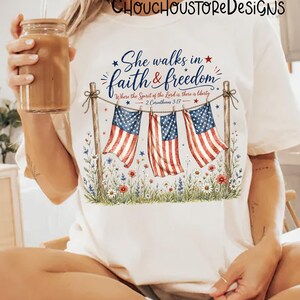 May include: Cream-colored t-shirt featuring a patriotic design. The design includes three American flags hanging from a rope, with the text "She walks in faith & freedom" and a Bible verse. Floral accents and stars complete the design.