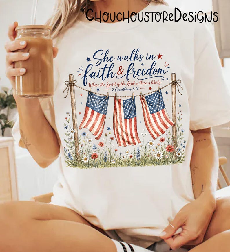 May include: Cream-colored t-shirt featuring a patriotic design. The design includes three American flags hanging from a rope, with the text "She walks in faith & freedom" and a Bible verse. Floral accents and stars complete the design.