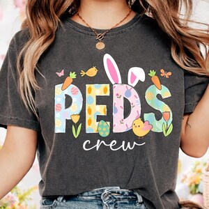 May include: Dark gray t-shirt with the text "PEDS crew" in colorful, Easter-themed lettering. The design includes bunny ears, carrots, chicks, butterflies, and floral elements, perfect for the spring season.