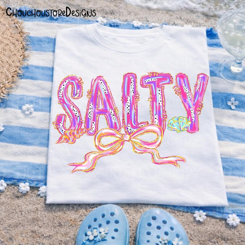 May include: White t-shirt with the word "SALTY" in large, colorful, bubble letters. The letters are pink, purple, and gold, with a bow at the bottom. The shirt is on a blue and white striped towel, with blue sandals at the bottom.