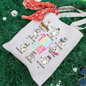 May include: A light-colored canvas tote bag with a colorful "Test Day" graphic. The design features cartoon ducks with encouraging phrases like "You got this" and "Keep your calm". The bag has long, off-white handles.