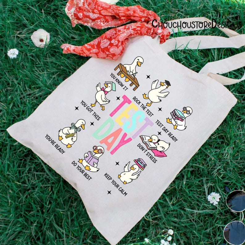 May include: A light-colored canvas tote bag with a colorful "Test Day" graphic. The design features cartoon ducks with encouraging phrases like "You got this" and "Keep your calm". The bag has long, off-white handles.