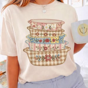 May include: A beige t-shirt featuring a stack of decorative casserole dishes. Each dish has a unique floral or patterned design, including pink, blue, and brown gingham. The dishes are stacked on top of each other.