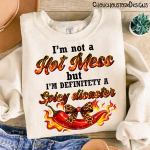 May include: Cream-colored sweatshirt with the text "I'm not a Hot Mess but I'm definitely a Spicy disaster." A red chili pepper with a leopard print bow and flames is featured below the text. The sweatshirt is folded.