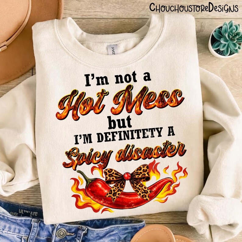 May include: Cream-colored sweatshirt with the text "I'm not a Hot Mess but I'm definitely a Spicy disaster." A red chili pepper with a leopard print bow and flames is featured below the text. The sweatshirt is folded.