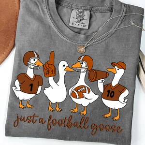 May include: Gray t-shirt with a graphic of four white geese in football gear. The geese wear helmets, jerseys, and hold a football and megaphone. The text "just a football goose" is below the geese.