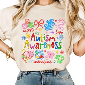 May include: Beige t-shirt with a colorful Autism Awareness design. The design includes the words "Autism Awareness" in red and yellow, along with various symbols like puzzle pieces, a ribbon, and a brain. Other words include "accept", "love", and "understand".