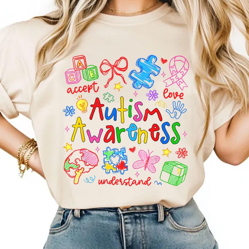 May include: Beige t-shirt with a colorful Autism Awareness design. The design includes the words "Autism Awareness" in red and yellow, along with various symbols like puzzle pieces, a ribbon, and a brain. Other words include "accept", "love", and "understand".