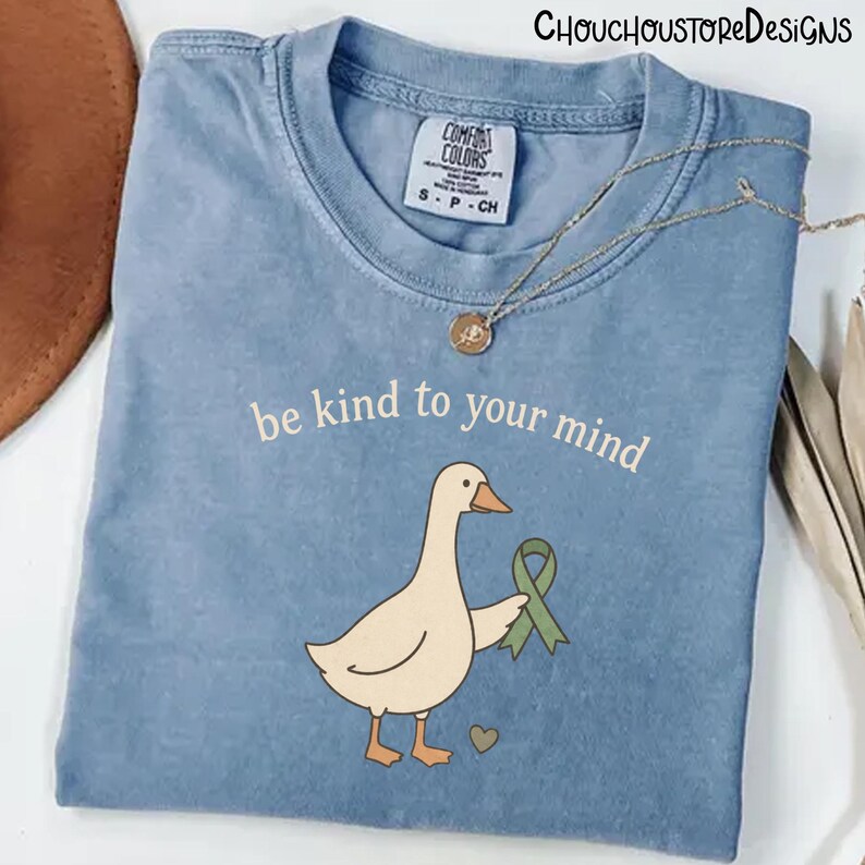 May include: A light blue Comfort Colors t-shirt with the phrase "be kind to your mind" and a cartoon goose holding a green ribbon. A gold necklace is draped over the shirt.