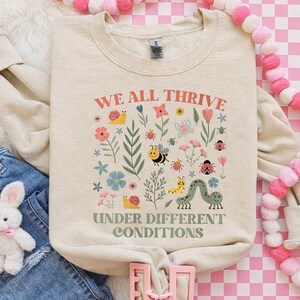 Peut inclure: Sweat-shirt cr&egrave;me avec le texte "WE ALL THRIVE" et "UNDER DIFFERENT CONDITIONS" en rouge et vert. Le motif comprend des illustrations color&eacute;es de fleurs, d'insectes et d'un escargot. Le sweat-shirt est pos&eacute; &agrave; plat.