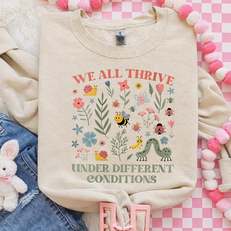 Peut inclure: Sweat-shirt cr&egrave;me avec le texte "WE ALL THRIVE" et "UNDER DIFFERENT CONDITIONS" en rouge et vert. Le motif comprend des illustrations color&eacute;es de fleurs, d'insectes et d'un escargot. Le sweat-shirt est pos&eacute; &agrave; plat.
