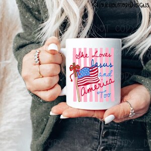 May include: White ceramic mug with the text "She Loves Jesus and America Too" in red and blue script. The mug features a graphic of the American flag on a wooden pole with a red bow, set against a pink striped background.