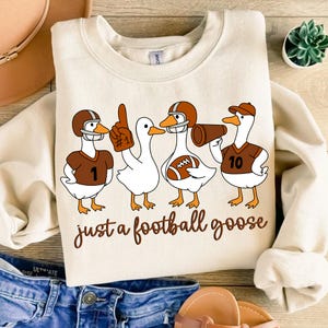 May include: Cream-colored sweatshirt featuring a whimsical design of four cartoon geese dressed as football players. The geese wear helmets and jerseys with numbers, and one holds a megaphone. The text "just a football goose" is written in a brown script.