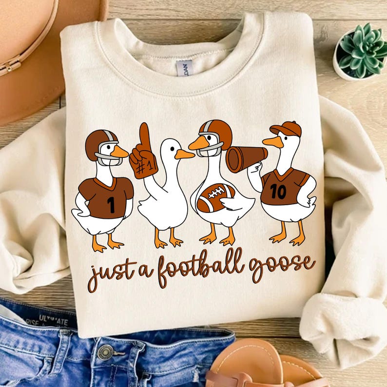 May include: Cream-colored sweatshirt featuring a whimsical design of four cartoon geese dressed as football players. The geese wear helmets and jerseys with numbers, and one holds a megaphone. The text "just a football goose" is written in a brown script.